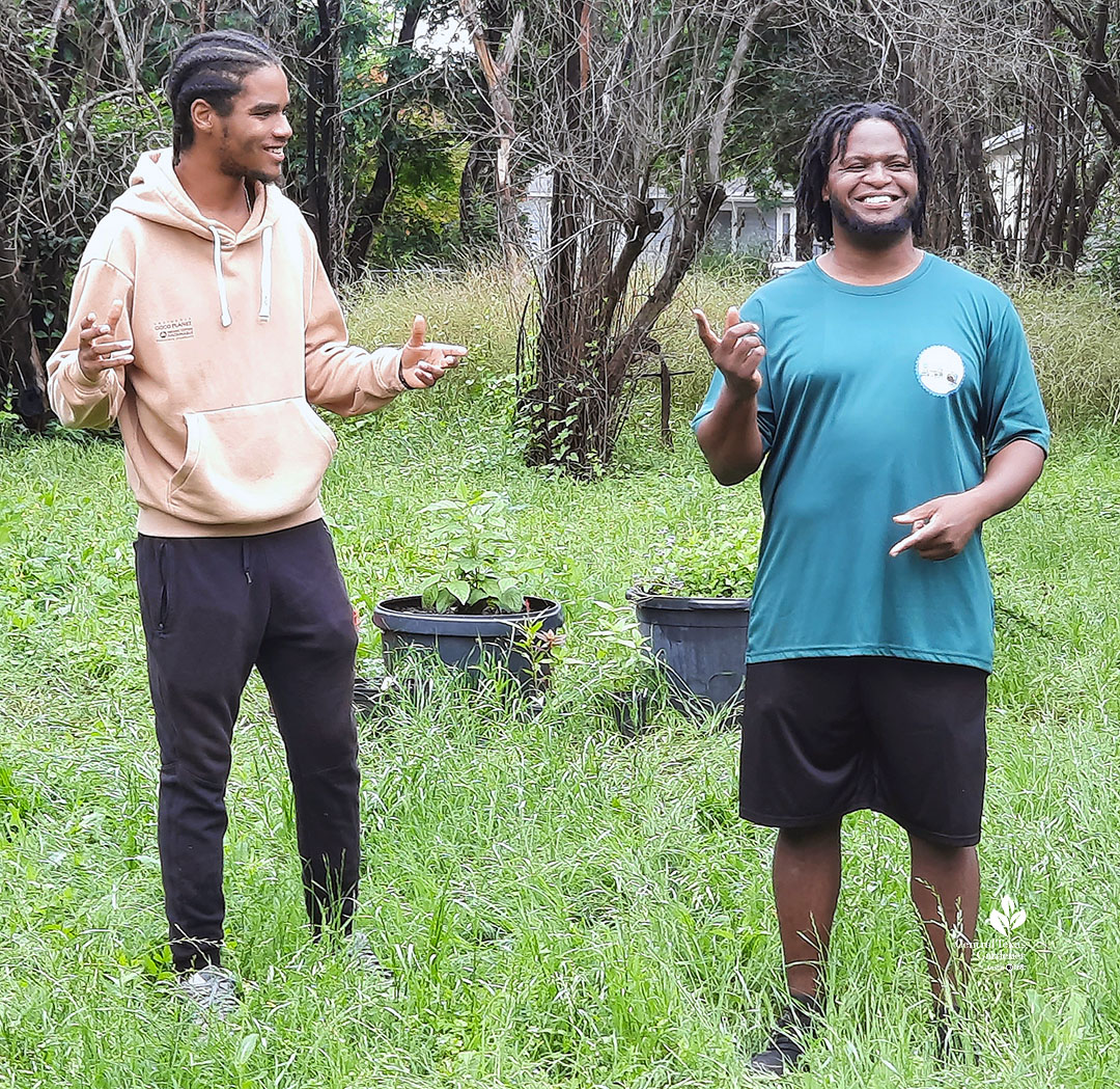 two young men smiling and gesturing to each other in field with grasses and trees 