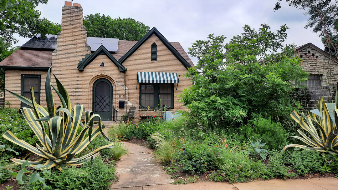 brick house with gabled roof and flowers, agaves, and small tree in front 