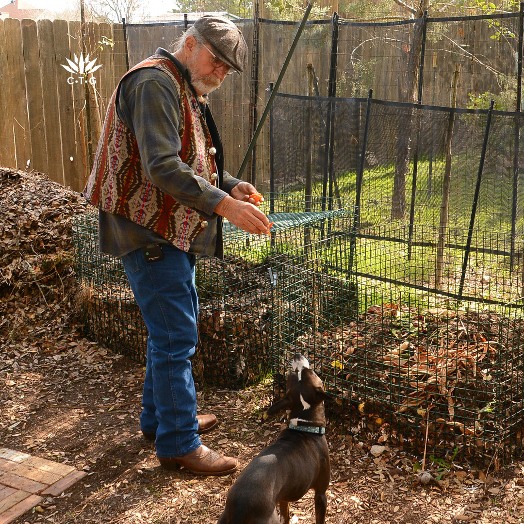 man adding vegetable scraps to compost pile and smiling at his dog 