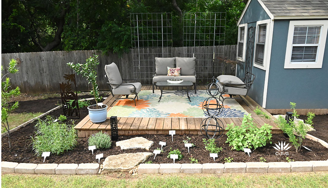 new garden beds in front of patio deck with furniture and a rug. A cattle panel trellis frames the back against a fence. 
