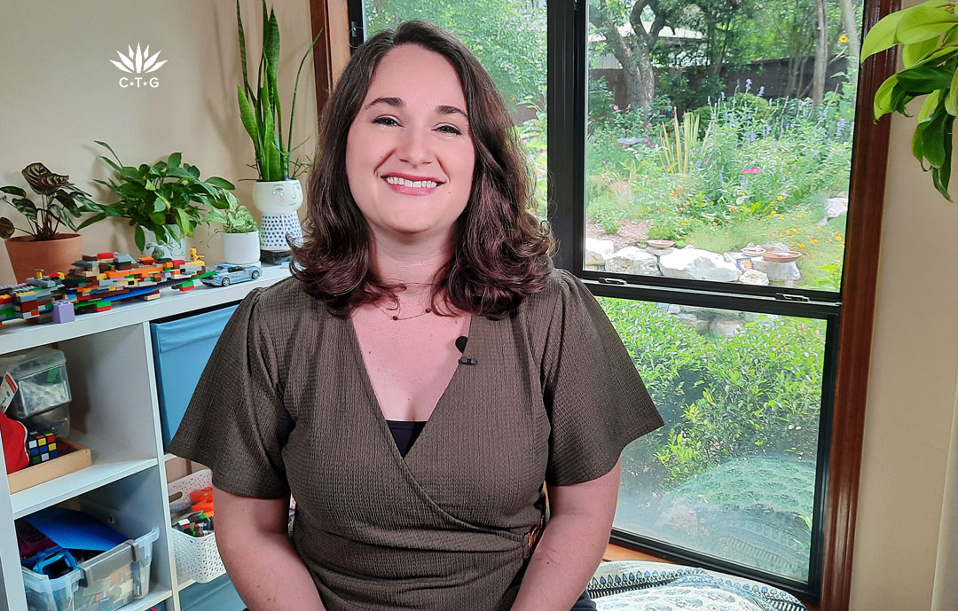 smiling woman sitting inside next to a bookcase with window and garden view behind her