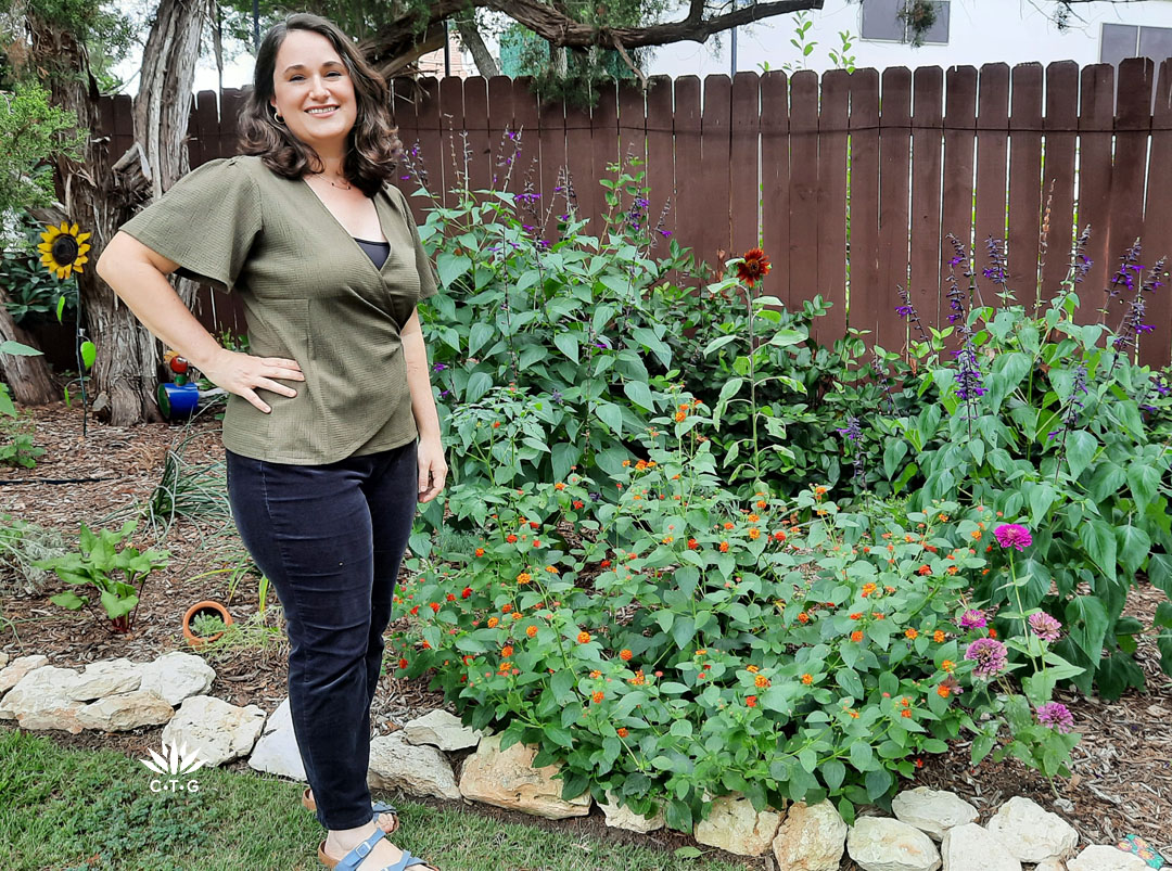 smiling woman next to fence garden filled with orange-flowered lantana and deep blue  flowered salvia 