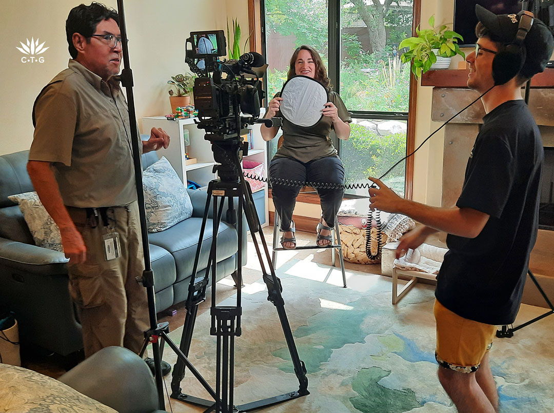 woman in living room holding white camera reflector in front of her; camera director laughing with audio person holding headphones to his ears 