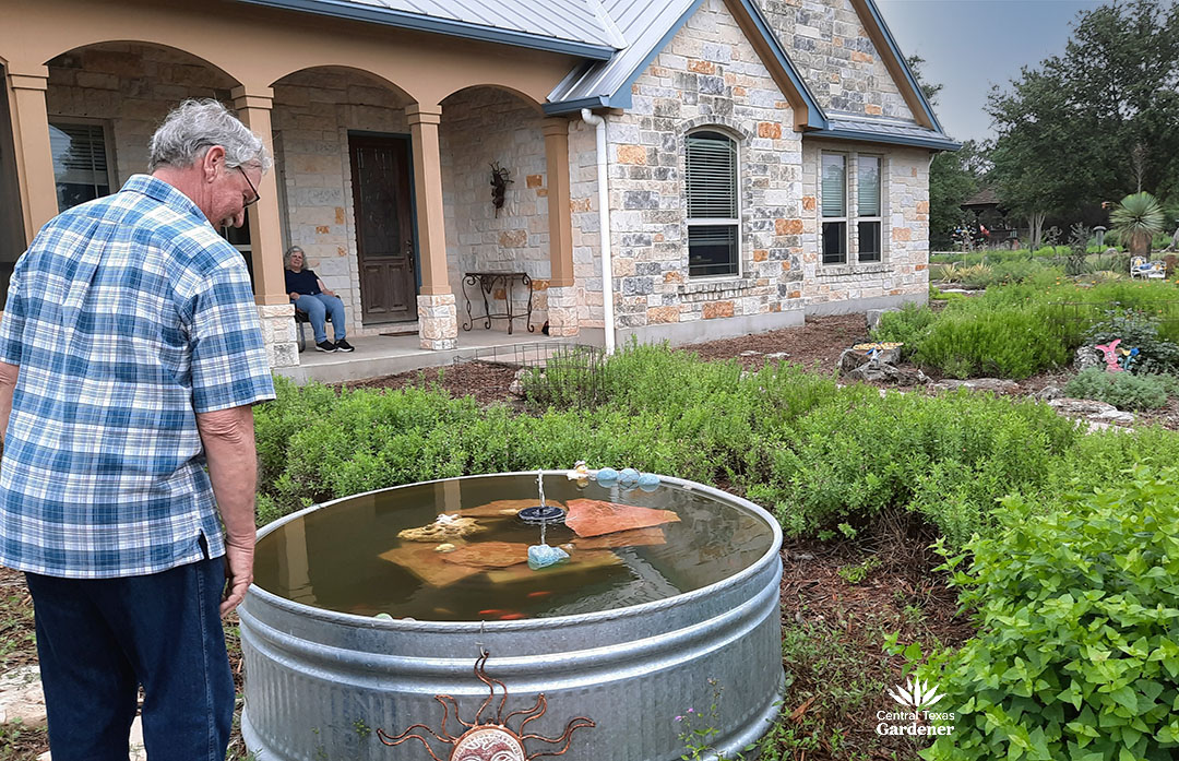 man in front of stock tank pond in front yard; woman watching from front porch